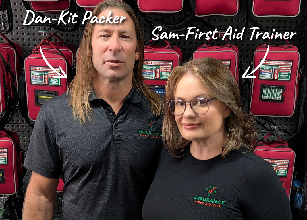 A man and a woman standing in front of a wall of red first aid kit pouches. White text labels identify them as 'Dan-Kit Packer' and 'Sam-First Aid Trainer'.